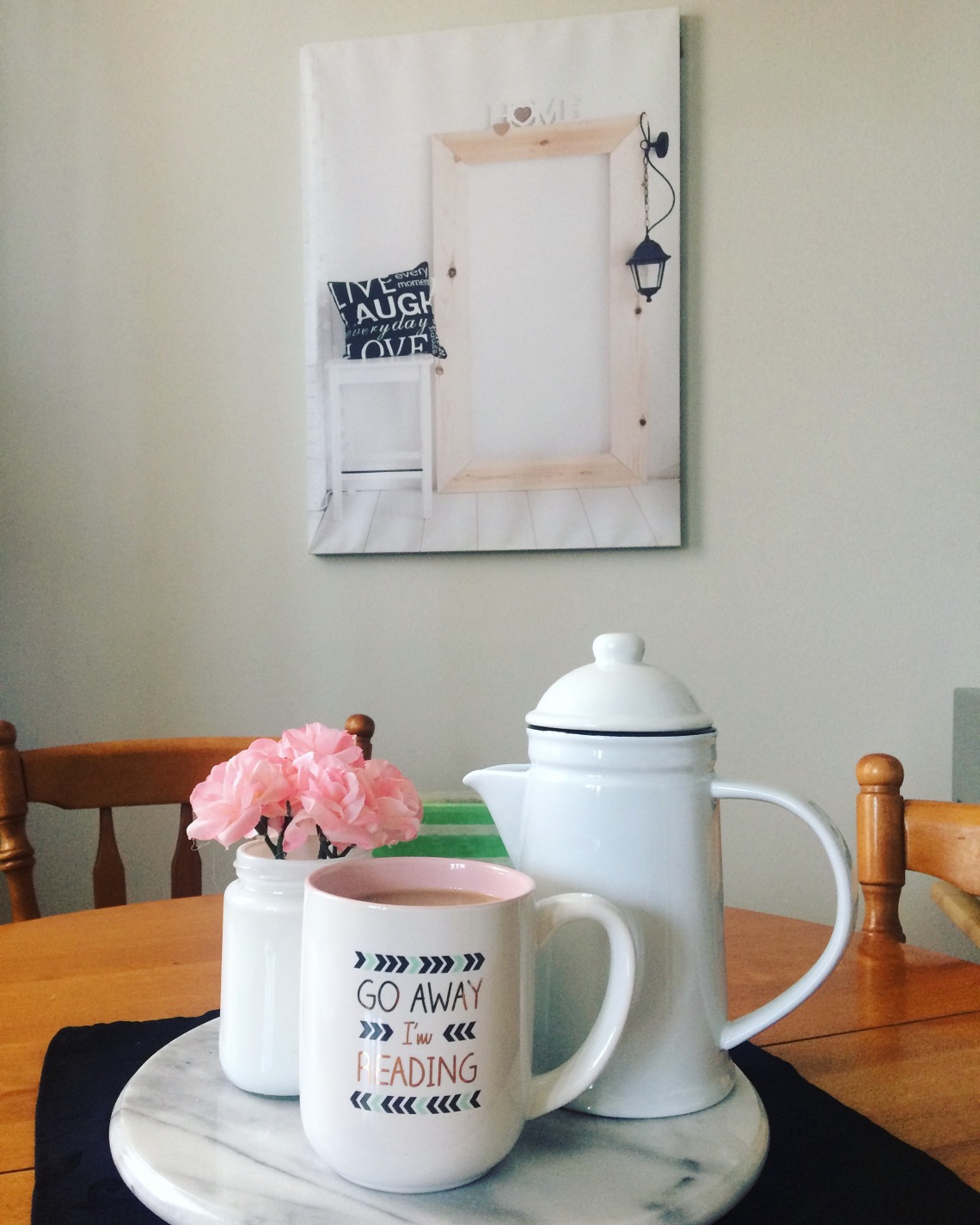 picture of Alyson's home: a picture on the wall of a door with a pillow on a white table that reads 'live, laugh, love' a coffee pot, mug and flowers on a wooden table. Coffee cup reads 'go away I'm reading'