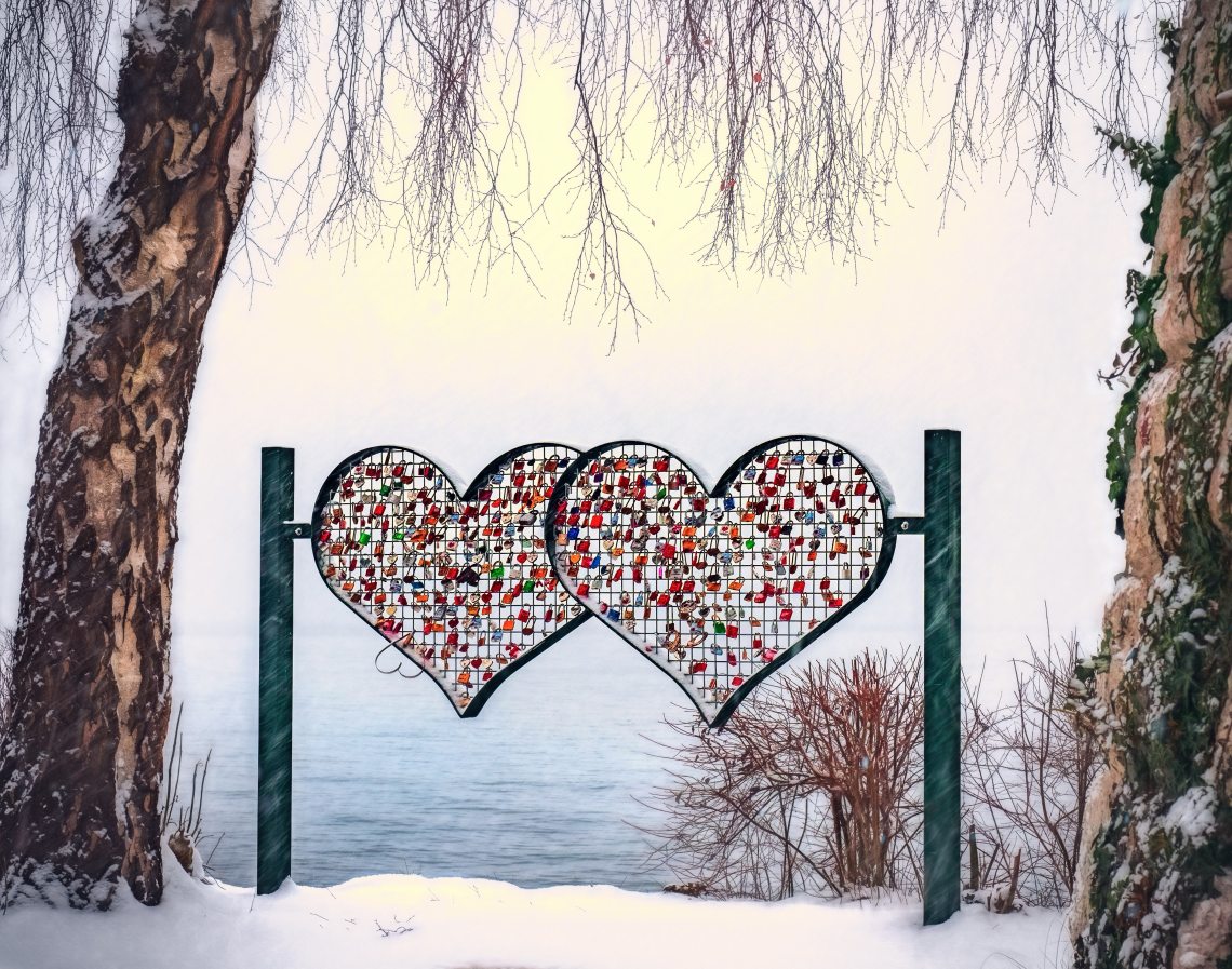 A metal sculpture of two hearts facing a lake in the winter with padlocks locked on the inside grid of both hearts