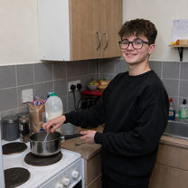 Young person preparing a meal independently in Oasis Care supported accommodation with staff support nearby.