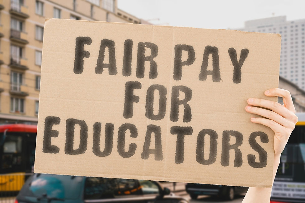 Hands holding a cardboard sign saying "FAIR PAY FOR EDUCATORS" in bold, black letters. City street and vehicles blurred in the background.