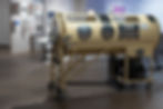 Beige wheeled hyperbaric chamber in a museum gallery, with metal portholes and display plaques on the wall.