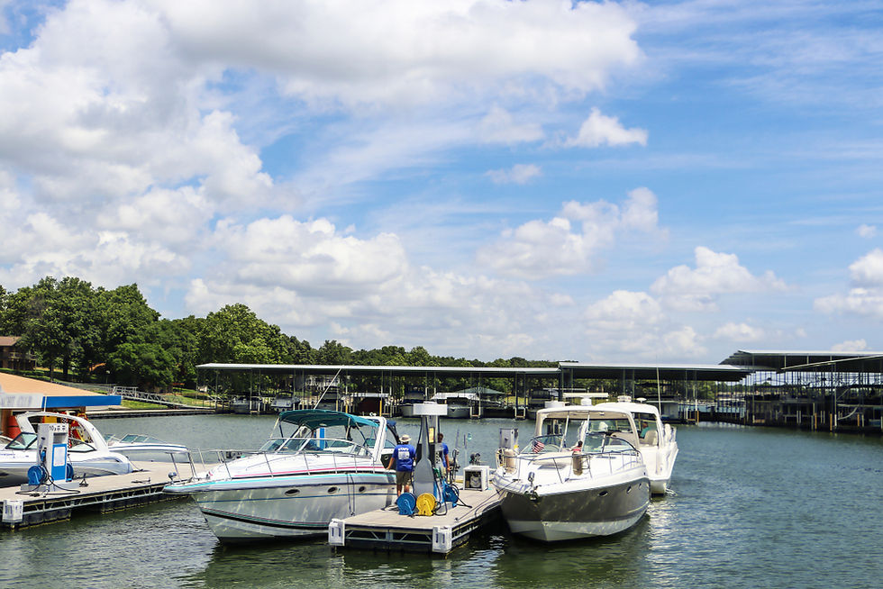 Boats docked at a marina on calm water under a bright blue, cloudy sky, with trees and covered slips in the background.