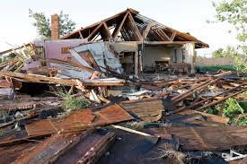 Collapsed house with wooden debris scattered in the foreground, amidst trees under a clear sky, indicating destruction or aftermath.