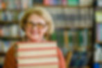 Smiling woman with glasses holding a stack of books in a library. Blurred bookshelves in the background. Warm and inviting mood.