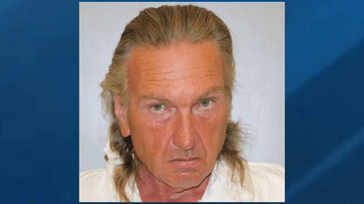 Stern-faced blond man in a mugshot-style portrait against a plain light background with a blue border.