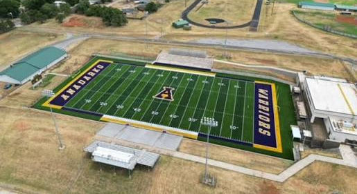 Aerial view of a green football field with yellow and purple borders, HILLVIEW and WARRIORS end zones, in a dry campus setting.