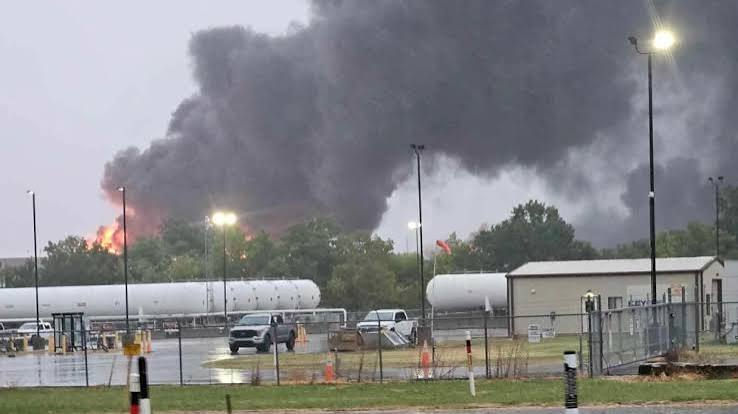 Large fire at an industrial site sends thick black smoke over parked trucks, tanks, and wet pavement at dusk.