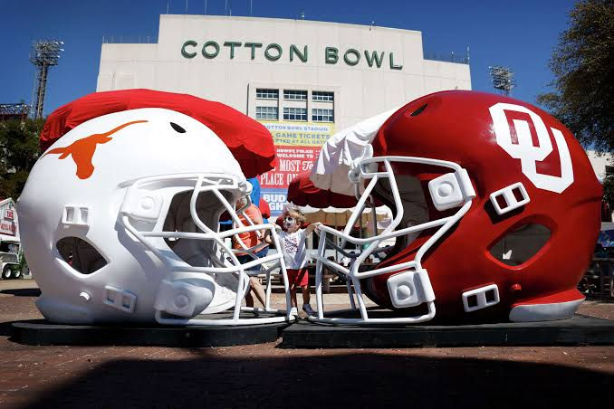 Giant Texas and Oklahoma football helmets face off outside Cotton Bowl stadium, with a person standing between them.