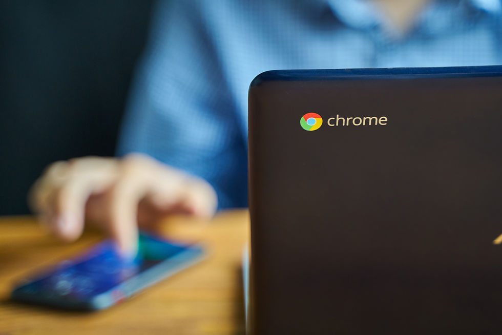Chromebook with "chrome" logo in focus, blurred hand using a smartphone, wooden table, and blue shirt in background, suggests multitasking.