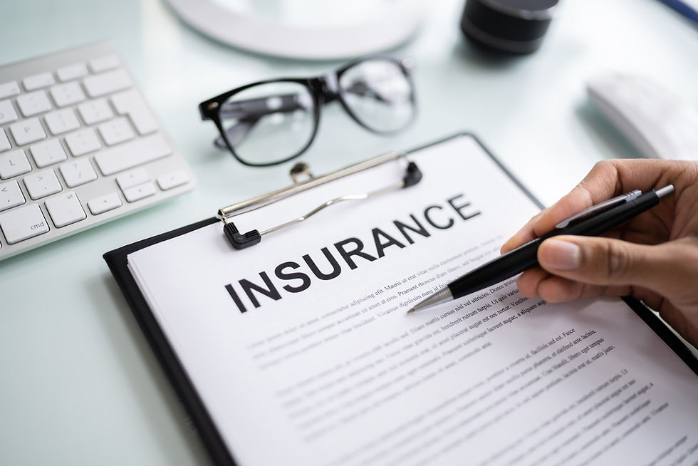 Hand holding pen over "INSURANCE" document on clipboard. Glasses, keyboard, and a lens cap are on the desk, conveying a professional setting.