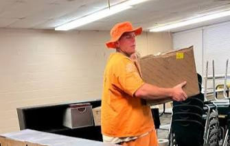Man in orange shirt and hat carries a cardboard box in a classroom with stacked chairs and desks.