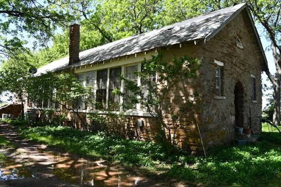 Stone cottage with steep roof and chimney among leafy trees, sunlit yard and shaded path.
