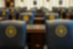 Rows of seats in a legislative chamber, featuring the Great Seal of the State of Oklahoma. The room is empty, with a formal atmosphere.