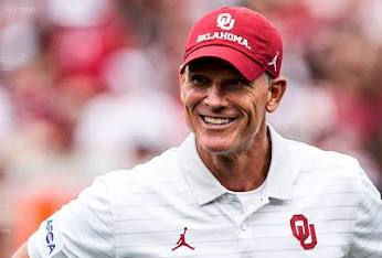 Smiling man in a red Oklahoma cap and white OU polo with Jordan logo, standing before a blurred crowd.