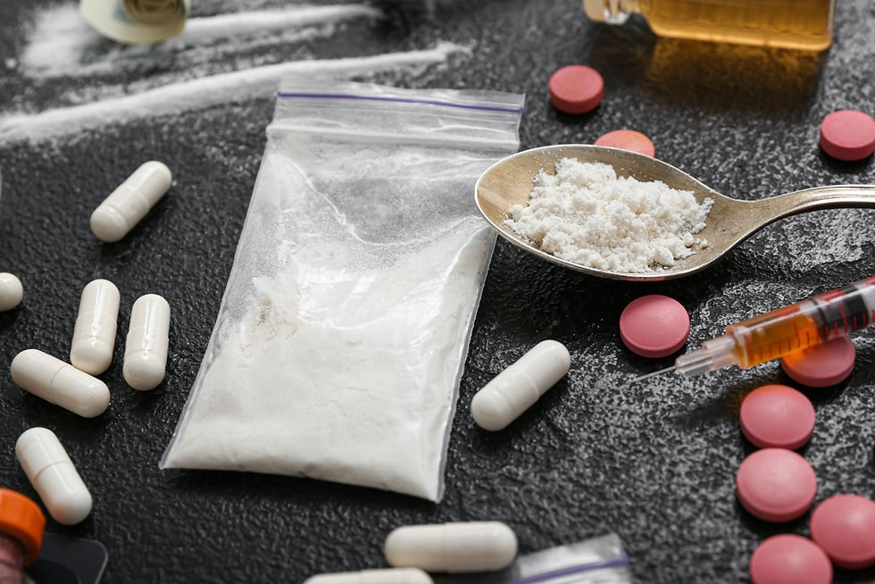 White powder in a plastic bag on a dark surface, with pills, a spoon, and a syringe, suggesting illegal drug use.
