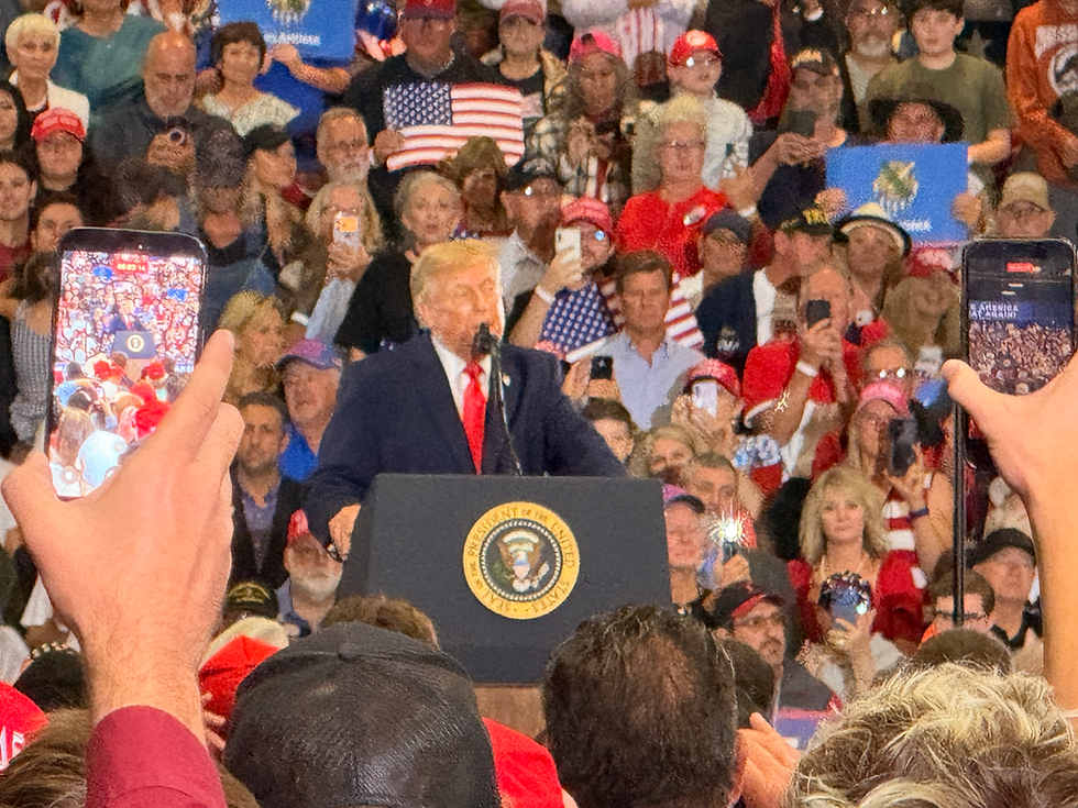 Donald Trump speaks at a podium with the presidential seal as a crowd films him, some holding U.S. flags, in a packed rally hall