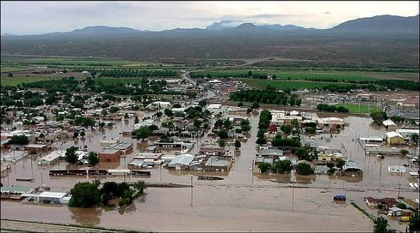 A flooded town with submerged streets and houses beneath muddy water, set in a valley with mountains under a cloudy sky.