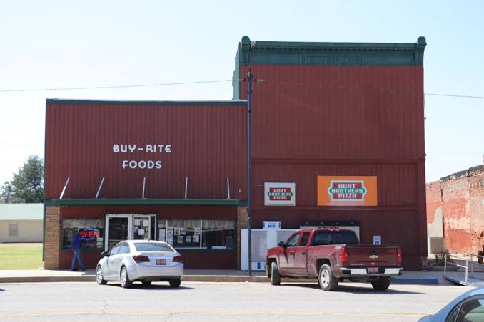 Buy-Rite Foods storefront with Hunt Brothers Pizza signs, cars parked out front, and a person near the open door on a sunny street.