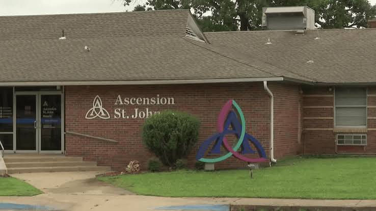 Brick Ascension St. John building with triquetra logo, glass entrance, and green lawn; calm exterior scene.