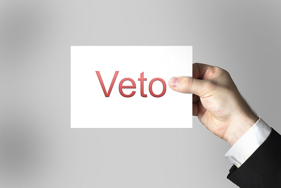 A hand in a suit holds a white card with "Veto" in red text on a gray background. The mood implies a formal decision or rejection.