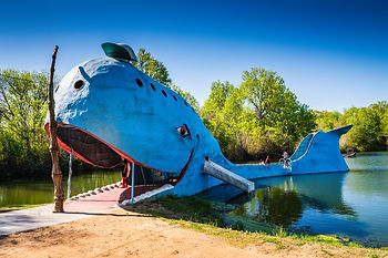 Giant blue whale-shaped structure by a lake, with people on its deck in a sunny green park under a clear blue sky.