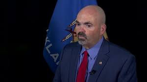 Bald man in a navy suit and red tie speaks before a blue flag, looking serious.