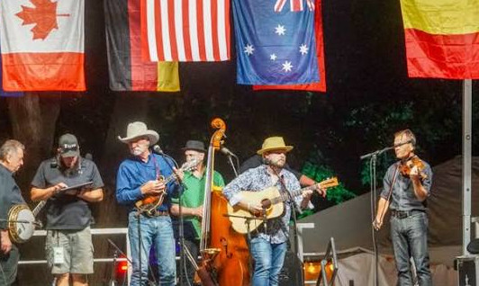 Bluegrass band performs on outdoor stage beneath hanging flags, with microphones, banjo, guitar, fiddle and upright bass; Ramm Tucker watermark
