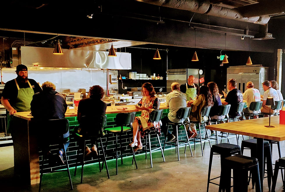 People seated at a bar in a dimly lit restaurant with dark decor, talking to chefs in green aprons. Mood is relaxed and social.