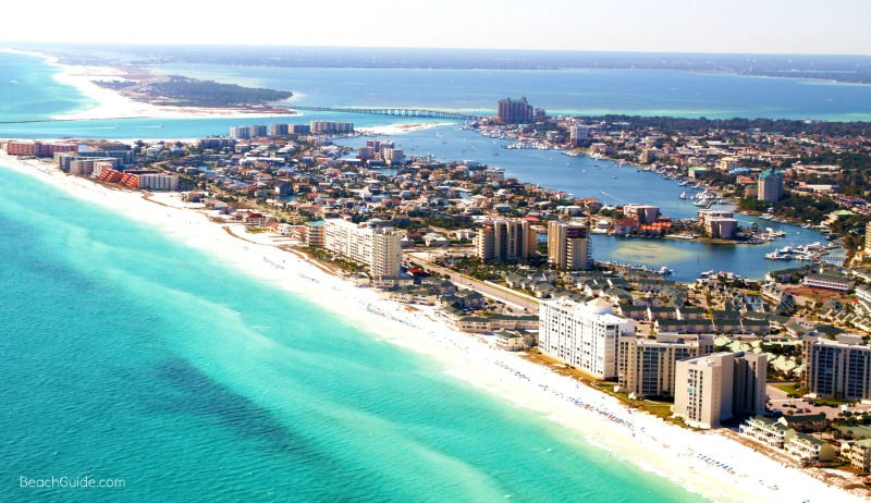 Aerial view of Destin, Florida with turquoise ocean, white sandy beaches, and numerous high-rise buildings. Calm, sunny day with a clear sky.