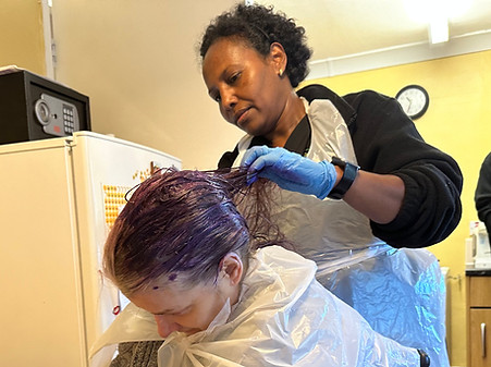 two women.one standing, one sitting.both wearing aprons.putting purple hair dye in hair.woman standing helping.in the kitchen