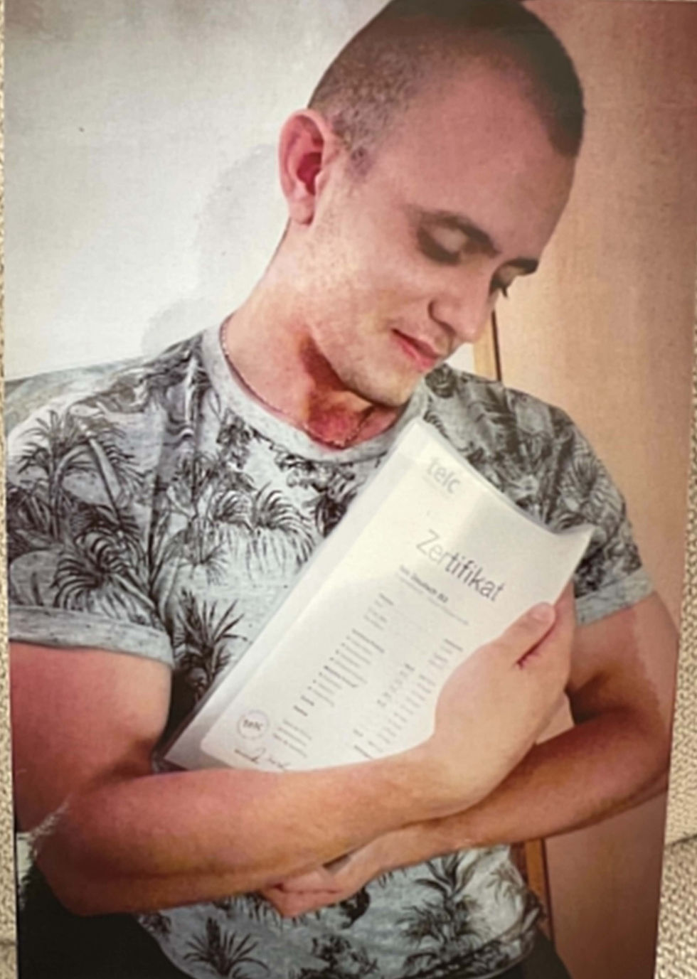 Alex Powerbilding in a patterned shirt hugs a certificate labeled "Zertifikat" with a content expression, set against a plain indoor background in Berlin, Germany