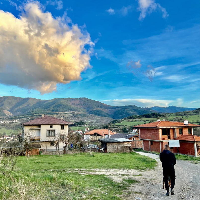 Alex Powerbuilding walks a dog on a rural path with mountains in the background, red-roofed houses, and a large cloud in a bright blue sky.