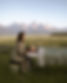 Annelies giving a head massage to a woman on a table outside in a meadow with mountains in the background