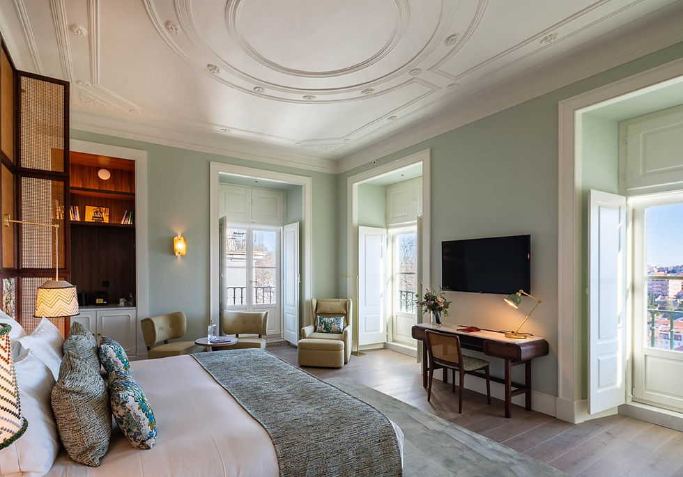 Elegant bedroom with a cozy bed, pillows, desk, TV, and armchairs. Bright sunlight streams through large windows, creating a serene ambiance.