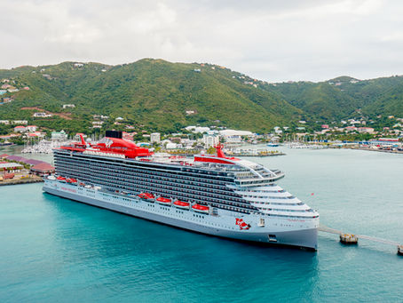 Cruise ship docked at a scenic port with green hills and colorful waterfront buildings. Calm turquoise water and overcast skies.