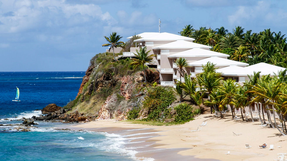 Cliffside resorts with white roofs overlook a sandy beach and turquoise sea. A sailboat on the water, palm trees in the background.