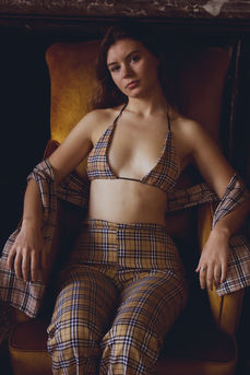 Young woman wearing a tartan bralette, trousers and shawl on a yellow velvet chair looking into the camera.