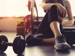 Kvinna sitter på gymgolvet och vilar med vattenflaska och hantel i förgrunden, i varmt ljus.