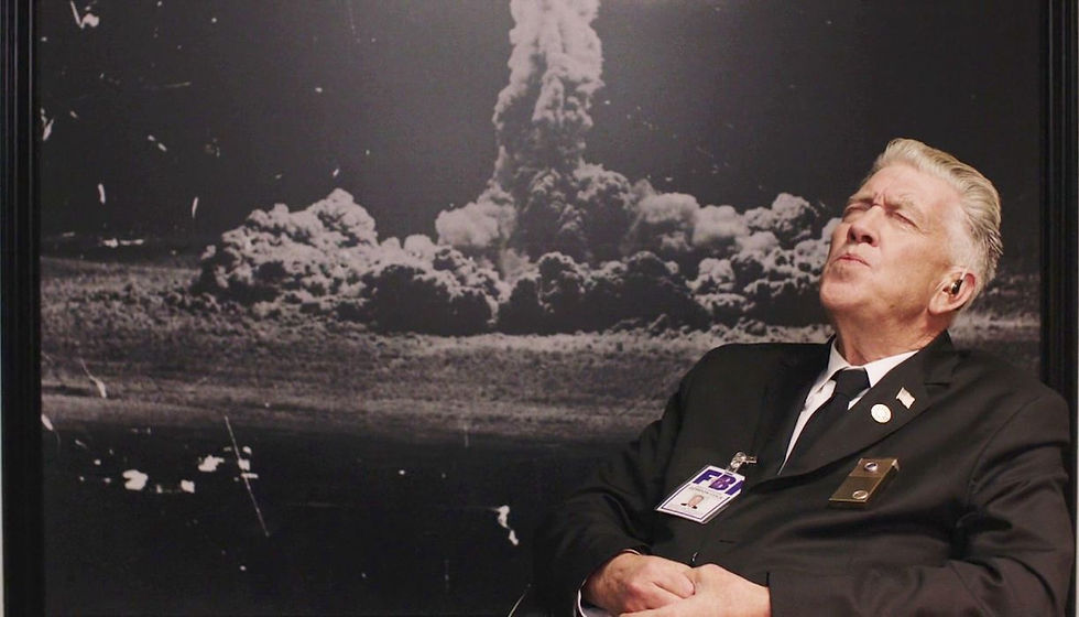 David Lynch in character as Gordon Cole in Twin Peaks whistling in a chair in front of a picture of a nuclear bomb.