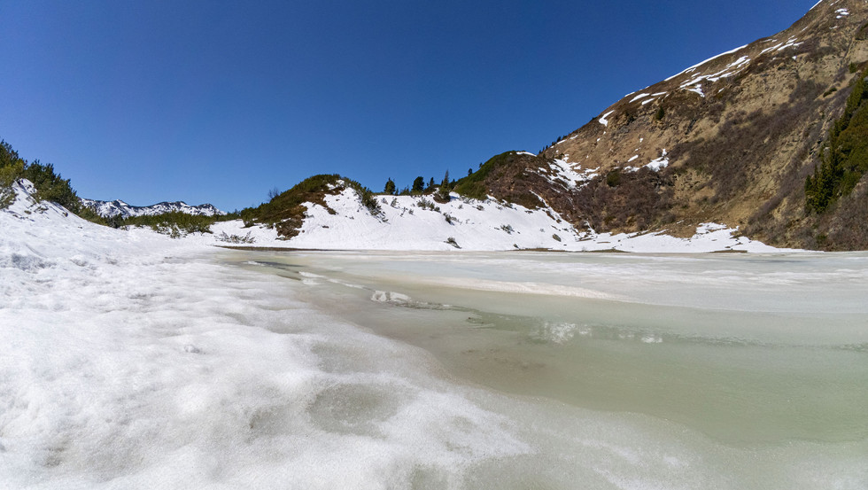 Ein schmelzender Plannersee inmitten der schneebedeckten Landschaft, mit Resten von Eis und schneebedeckten Hügeln im Hintergrund.