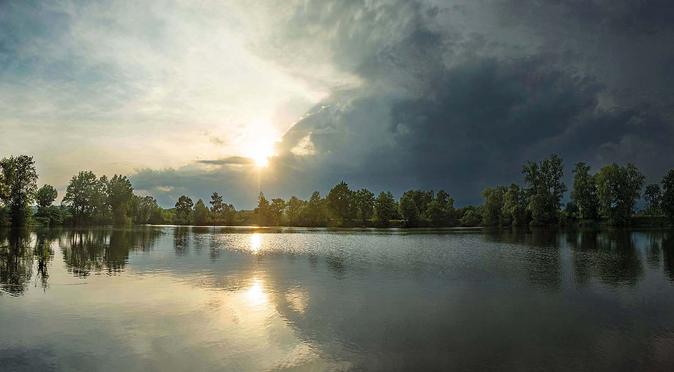 Dieses Panorama fängt die geladene Atmosphäre kurz vor dem Hagelschauer ein. Das Bild lebt vom extremen, diagonalen Kontrast: Während auf der linken Seite die tiefstehende Nachmittagssonne das Wasser und die Uferbäume in ein warmes, goldenes Licht taucht, schiebt sich von rechts eine massive, bedrohlich dunkle Gewitterfront ins Bild. Die spiegelglatte Wasseroberfläche reflektiert beide Welten gleichermaßen.