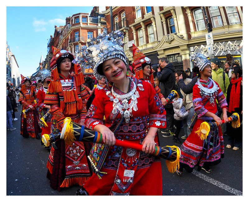 Parade with people in vibrant red, patterned costumes, and silver headdresses, smiling and holding colorful staffs. City street backdrop. Festive mood. Chinese New Year, London