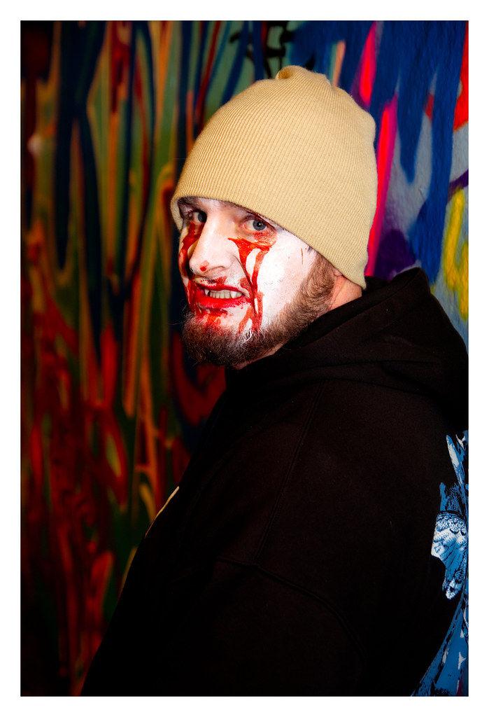 Bearded vampire man with a painted bloody face wearing a beige beanie, standing against a colorful graffiti wall. The expression is intense.
