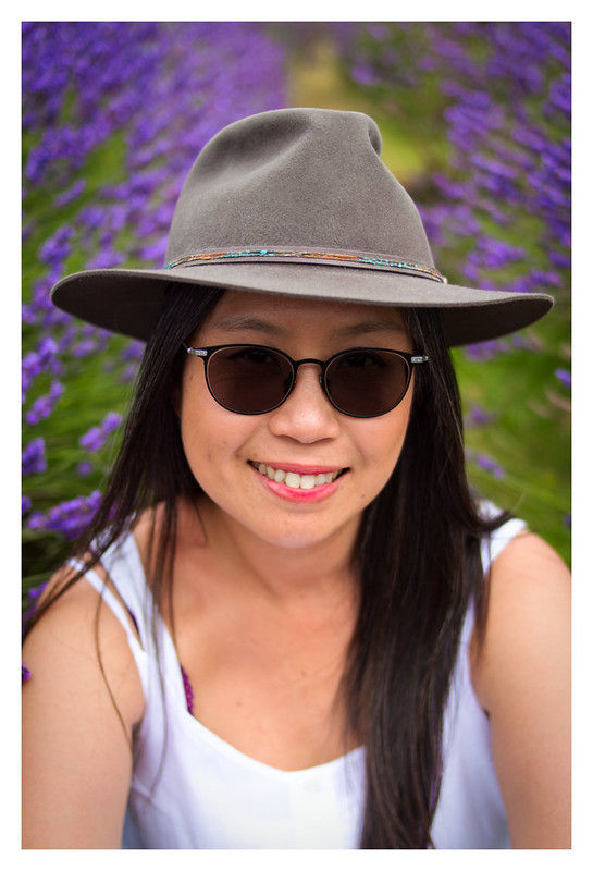A person in a hat and sunglasses smiles in a lavender field background. Wearing white with vibrant purple flowers, creates a cheerful mood.