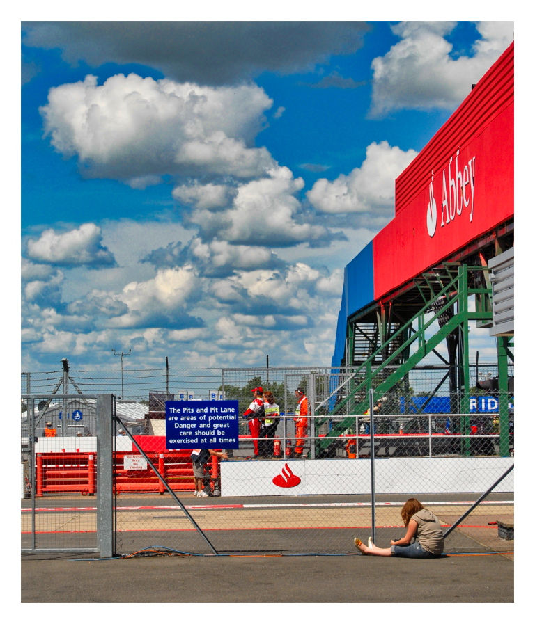 Race track scene with people in safety gear near pit lane, clouds above. A person sits alone on the ground. Abbey sign visible.