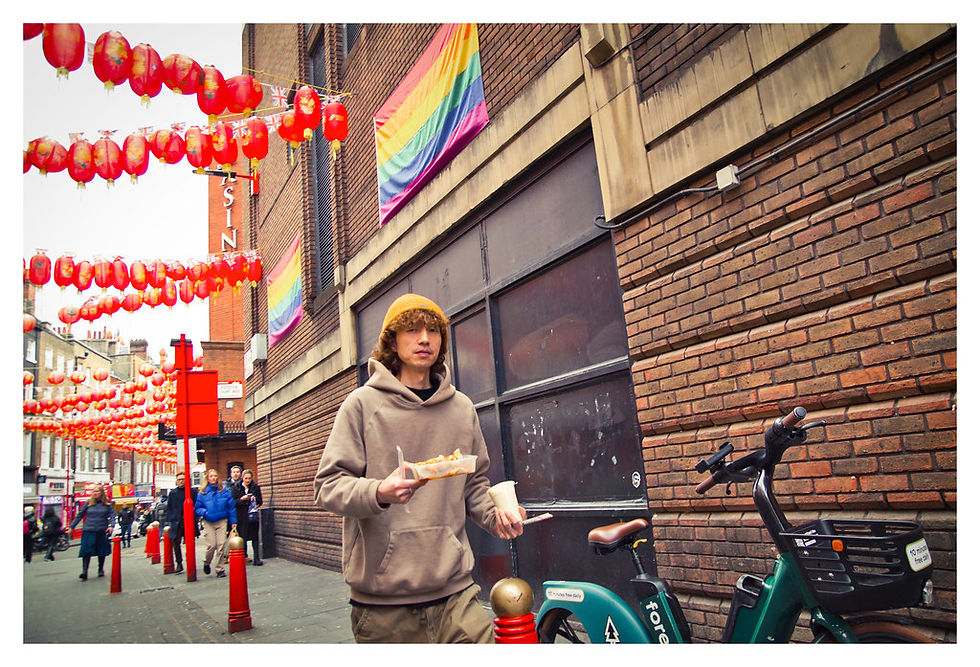 Person in a hoodie holds food and drink, walking past red lanterns and a rainbow flag in a city street. Bike parked by a brick wall.