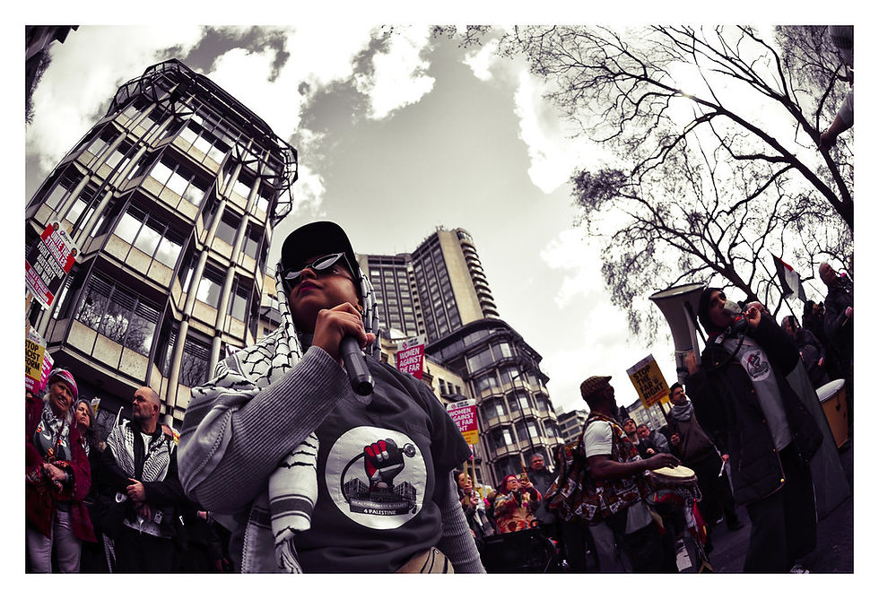 A city protest shows a person with a mic and others holding signs. Buildings and bare trees are in the background. The mood is determined. Together Alliance, London, March 2026
