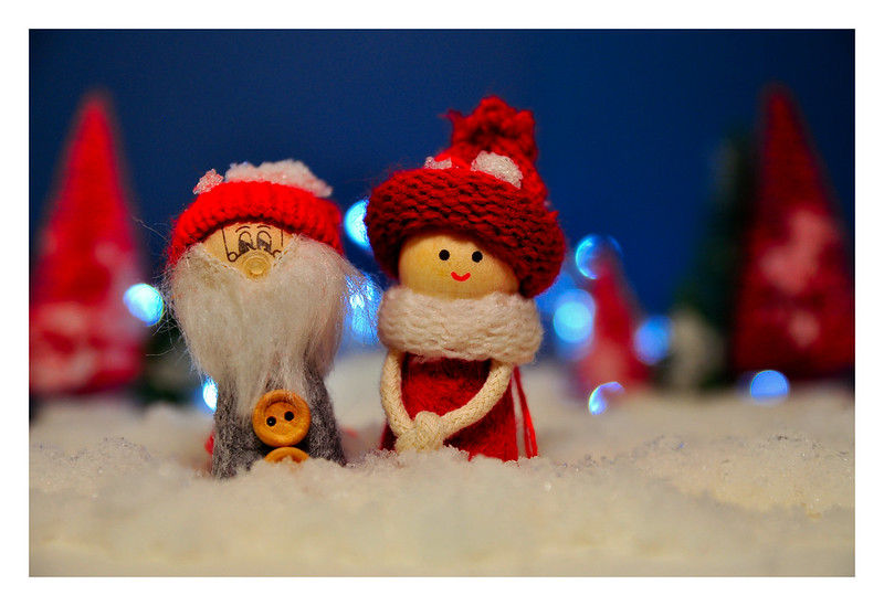 Two wooden figures, elf man and woman, in red knit hats and scarves, stand on snow. Blurred red trees and blue lights in the background.