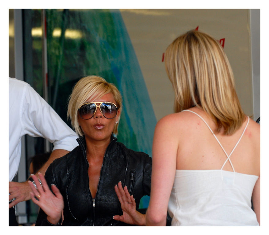 Posh Spice, Victoria Beckham in sunglasses and black jacket gestures to another woman in a white top. Indoor setting with a colorful abstract background.