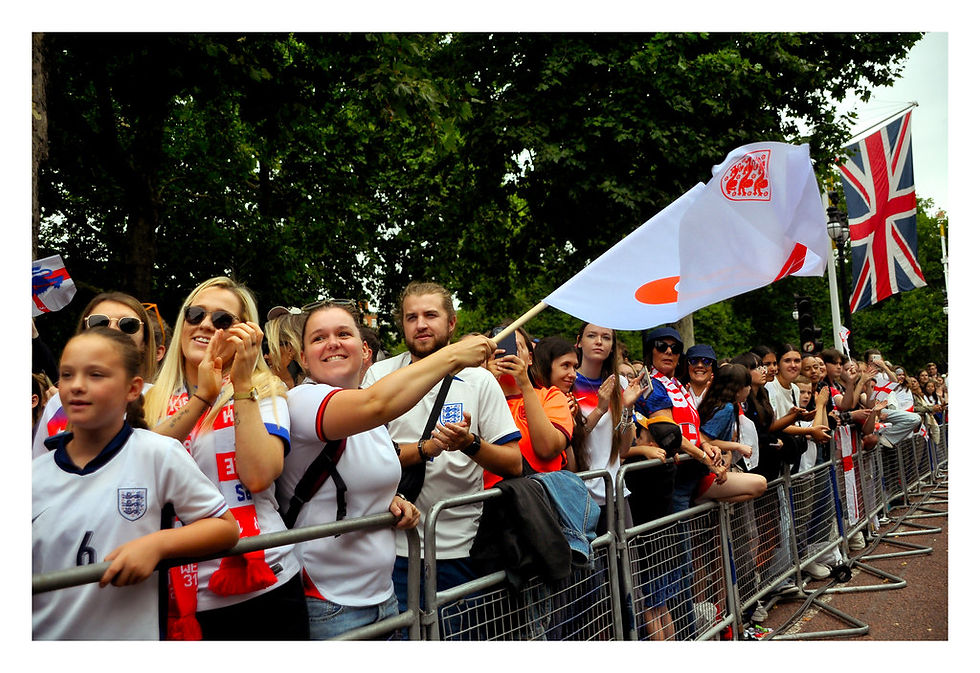 Crowd of fans in football jerseys and scarves, smiling and excited, waving flags. They're behind barriers with a Union Jack in the background. Celebrations of England Women's Euro 2025 win at the Victory Parade